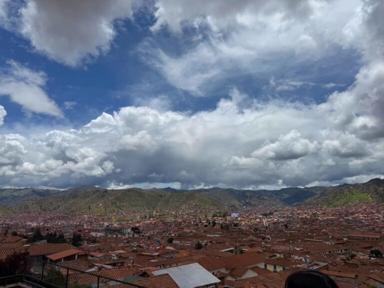 Red-tiled rooftops of Cusco beneath green hills and large clouds, seen from San Blas