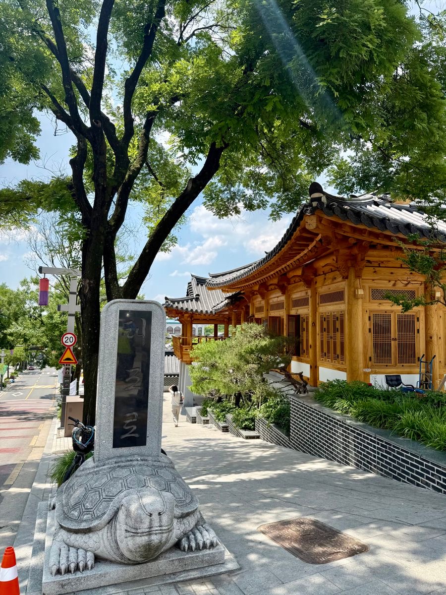 Traditional wooden building with tiled roofs beside a shaded street and stone turtle monument in Jeonju